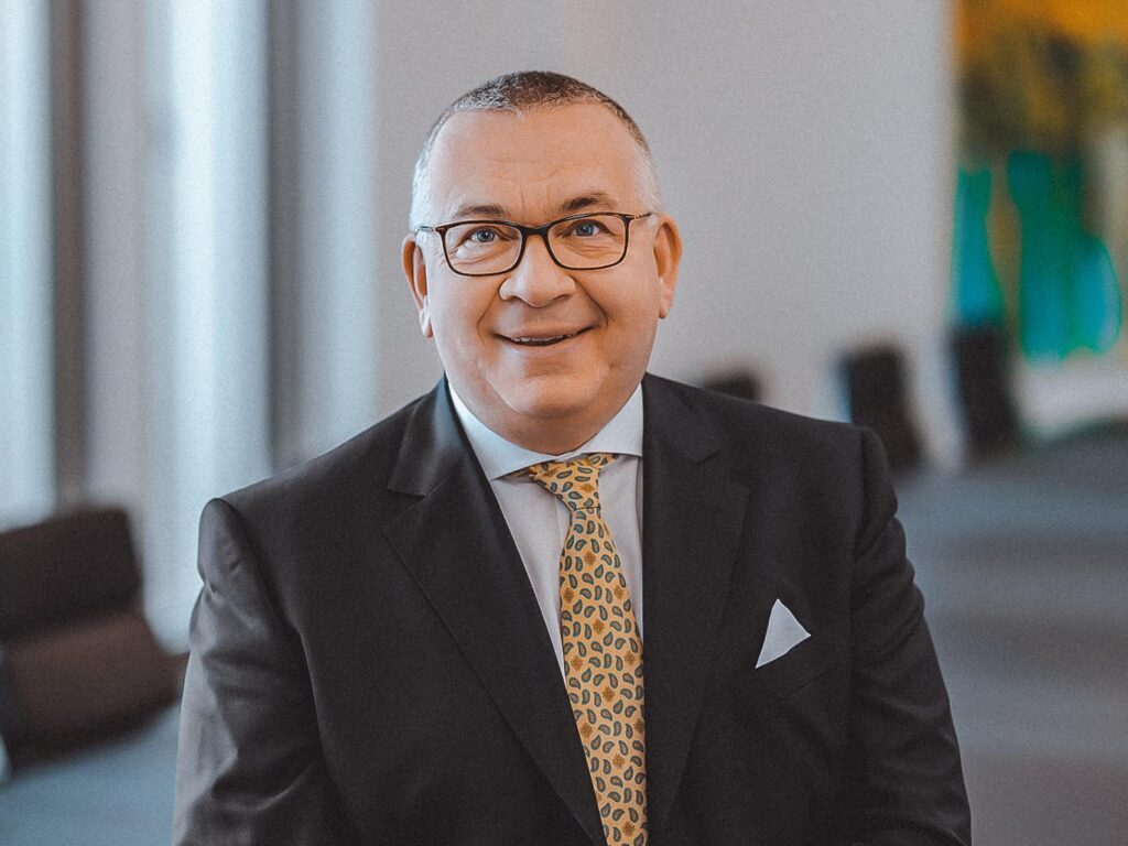 Das Bild zeigt den Aufsichtsratsvorsitzenden der Volksbank Kassel-Göttingen, Claus Winneknecht, in einem dunkelgrauen Anzug. Er trägt ein weißes Hemd und eine hellorange Krawatte sowie ein dazu passendes Brusttaschentuch in weiß. Winneknecht trägt eine Brille und kurz geschorene Haare und lächelt in die Kamera. Er steht in einem Raum mit einem, direkt hinter ihm sieht man unschwarf einen grauen Tisch sowie mehrere Bürostühle. Winneknecht sieht man nur von der Brust aufwärts.