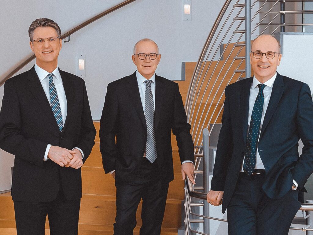 Der komplette Vorstand der Volksbank Kassel Göttingen steht vor einer Holztreppe, die nach ober führt. Es sind zu sehen, von links nach rechts: Vorstandsvorsitzender Wolfang Osse, Volker Stern und Hans-Christian Reuß