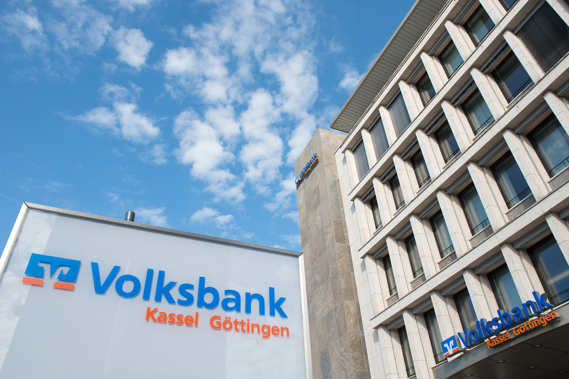 Das Bild zeigt die Hauptstelle der Volksbank Kassel Göttingen in Kassel. Links unten ist das Logo der Volksbank vor der Hauptstelle zu sehen. Rechts wiederum ist die Frontfassende des Gebäudes zu sehen.
