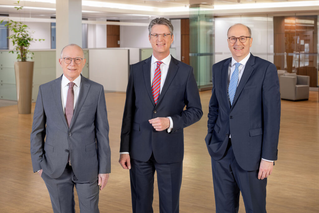 Der Vorstand der Volksbank Kassel Göttingen steht in einem Büro und schaut in die Kamera. Zu sehen sind, von links nach rechts, Volker Stern, Vorstandsvorsitzender Wolfang Osse sowie Hans-Christian Reuß. Alle drei tragen einen dunkelgrauen Anzug, nur Volker Stern einen Hellgrauen. Volker Stern trägt außerdem eine blau-orange gepunktete Krawatte, Wolfgang Osse wiederum eine rot-blau gestreifte Krawatte und Hans-Joachim Reuß eine blau-weiß gestreifte Krawatte.