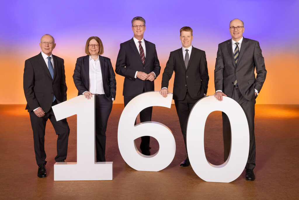 Das Bild zeigt den gesamten Vorstand der Volksbank Kassel Göttingen hinter einer großen, weißen Zahl, die "160" zeigt. Zusehen sind von links nach rechts: Volker Stern, Kerstin Orth, Wolfgang Osse, Thorsten Schartel und Hans-Christian Reuß. Alle tragen einen Anzug sowie ein weißes Hemd bzw. eine weiße Bluse. Dabei lächeln sie in die Kamera und stehen locker hinter der Zahl.