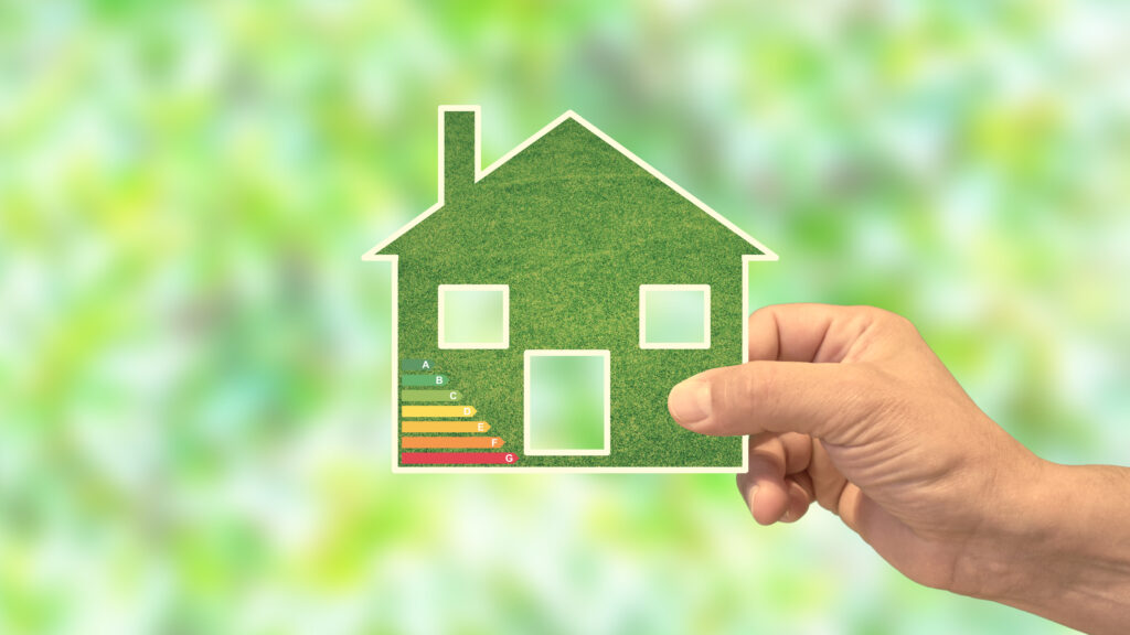Eine Hand hält ein umweltfreundliches grünes Haus mit Energieeffizienzbewertung. Im Hintergrund sind unscharfe Blätter zu sehen.