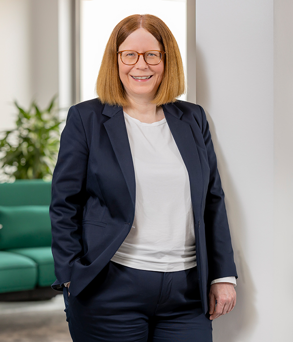 Auf dem Bild ist das Vorstandsmitglied Kerstin Orth zu sehen. Sie ist leicht an einer Wand angelehnt, im Hintergrund ist ein Büro zu sehen. Sie trägt ihr dunkelblaues Sakko offen. Die rechte Hand steckt in der rechten Hostentasche, die linke Hand hängt locker an der linken Seite runter.