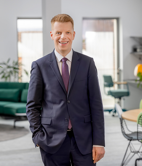 Im Bild steht Vorstandsmitglied Thorsten Schartel in einem Büro uns lächelt dabei in die Kamera. Er trägt sein Sakko geschlossen, seine orange-blau karierte Krawatte ist dennoch zu sehen. Seine rechte Hand steckt in der Hostentasche, seine linke Hand hängt locker an der Seite herunter.
