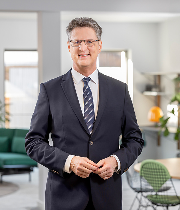 Auf dem Bild ist Wolfang Osse, Vorstandsvorsitzender der Volksbank Kassel-Göttingen in einem dunkelgrauen Anzug zu sehen. Er trägt ein weißes Hemd und eine dunkel- und hellblau gestreifte Krawatte. Seine Hände hat er vor sich auf dem Bauch verschlossen. Im Hintergrund ist ein Büro zu sehen.