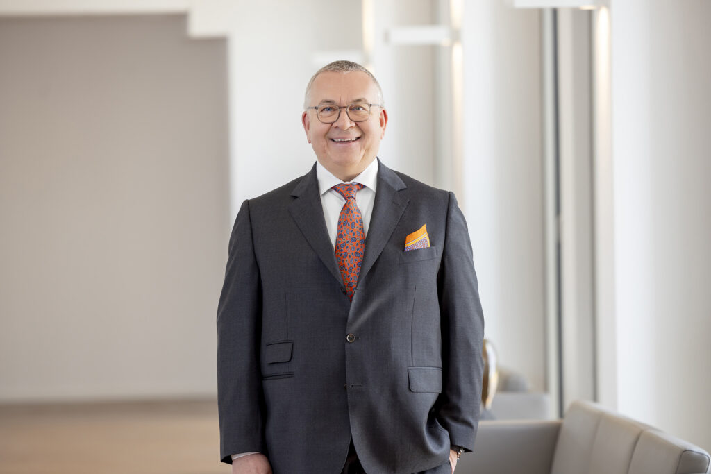 Das Bild zeigt den Aufsichtsratsvorsitzenden der Volksbank Kassel-Göttingen, Claus Winneknecht, in einem dunkelgrauen Anzug. Er trägt ein weißes Hemd und eine orange-blaue Krawatte sowie ein dazu passendes Brusttaschentuch. Sein linker Arm steckt in der Hosentasche, der rechte Arm hängt locker an der Seite herunter. Winneknecht trägt eine Brille und kurz geschorene Haare und lächelt in die Kamera. Er steht in einem Raum mit einem hellen Holzboden und einer weißen Wand.