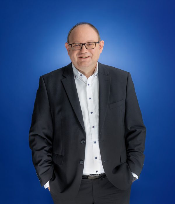 Das Bild zeigt Lars Bering. Er hat seine Hände in den Hosentaschen gesteckt, steht aber locker vor einem blauen Hintergrund und schaut lächelnd in die Kamera.