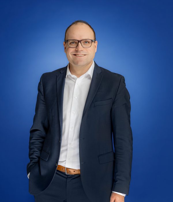 Das Bild zeigt Stefan Bömeke, wie er lächelnd, vor einem blauen Hintergrund, locker stehend in die Kamera lächelt.