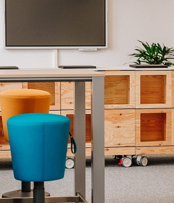 Das Bild zeigt einen modern eingerichteten Raum. Zu sehen ist im Hintergrund auf einer weißen Wand ein schwarzer Bildschirm. Darunter ist ein Holz-Sideboard zu sehen, auf dem eine Pflanze steht. vor dem Sideboard wiederum stehen ein Tisch sowie vier Hocker - je zwei in blauer und orangener Farbe. Der Boden ist ein grauer Teppichboden.