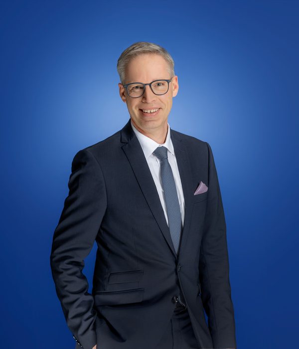 Das Bild zeigt Jochen Diemer. Er lächelt in die Kamera und steht dabei vor einem blauen Hintergrund. Er steht geradlinig da und lächelt in die Kamera.