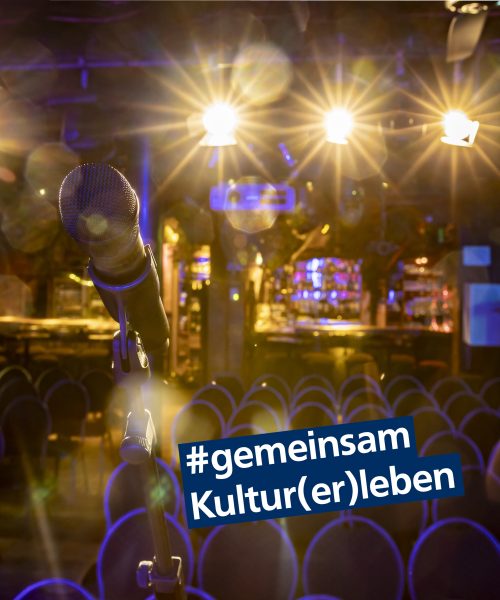 Das Bild zeigt einen Saal, von der Bühne aus fotografiert. Im Vordergrund ist ein Mikrofon zu sehen, davor mehrere Stühle und im Hintergrund eine Bar. Ober der Bar sind drei Scheinwerfer angebracht, die in die Kamera leuchten und so Blendeneffekte entstehen. Im unteren Drittel des Bildes steht außerdem "#gemeinsam Kultur(er)leben"