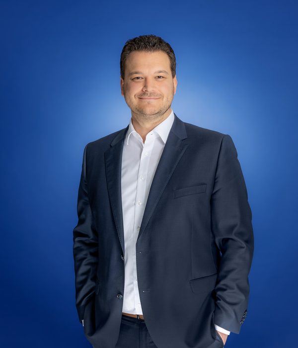 Das Bild zeigt Bastian Hartje. Er steht locker und entspannt vor einem blauen Hintergrund und schaut dabei amüsiert in die Kamera.