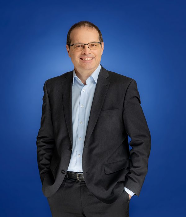 Das Bild zeigt Ralf Hildebrand im Anzug. Er lächelt in die Kamera und steht vor einem blauen Hintergrund. Er trägt einen Anzug.