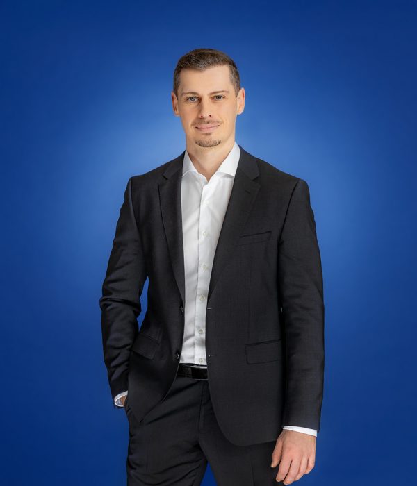 Das Bild zeigt Sven Mader, wie er locker vor einem blauen Hintergrund steht und in die Kamera schaut.