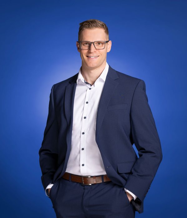 Das Bild zeigt Dennis Möller, wie er locker vor einem blauen Hintergrund in die Kamera lächelt.