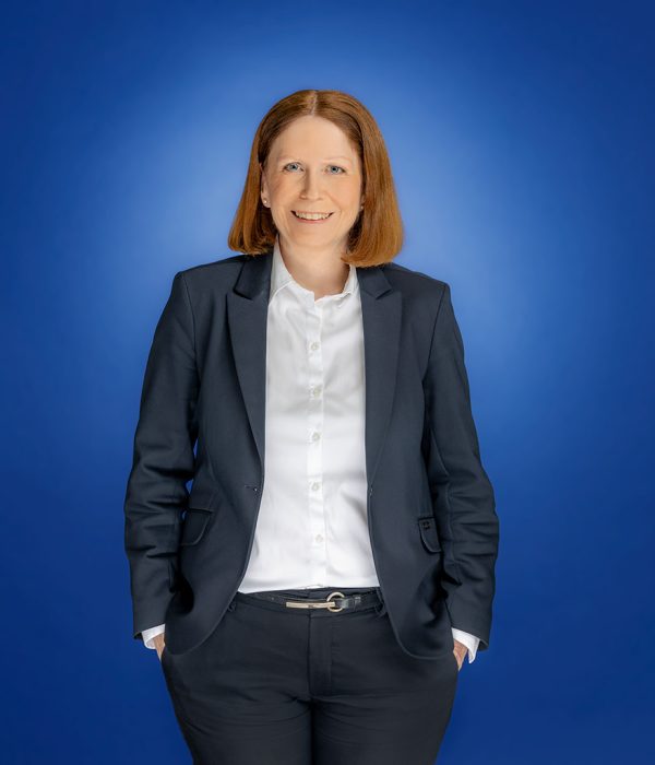 Das Bild zeigt Kerstin Orth, die in die Kamera lächelt. Ihre beiden Hände stecken in der Hosentasche und sie trägt ihr Sakko offen, sodass man ihr weißes Hemd sieht.