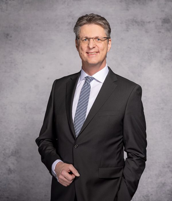 Im Bild ist der Vorstandsvorsitzende der Volksbank Kassel Göttingen Wolfgang Osse vor einem grauen Hintergrund zu sehen. Er trägt einen schwarzen Anzug, eine hellgrau-weiß gestreifte Krawatte sowie ein weißes Hemd. Sein Sakko ist zugeknöpft. Seine linke Hand steckt in der Hostentasche, sein rechter Arm hängt angewinkelt vor seinem Bau. Er lächelt in die Kamera.