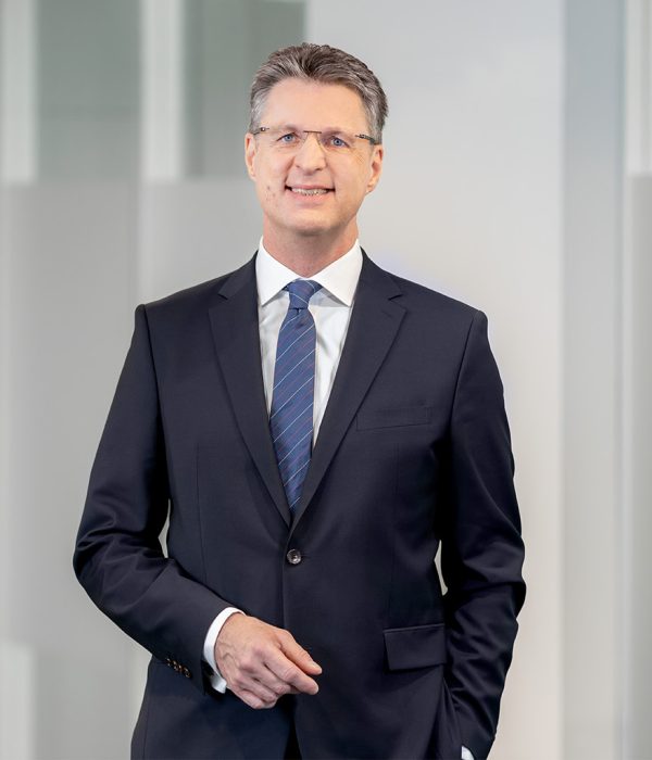 Auf dem Bild ist Wolfang Osse, Vorstandsvorsitzender der Volksbank Kassel-Göttingen in einem dunkelgrauen Anzug zu sehen. Er trägt ein weißes Hemd und eine dunkelblaue Krawatte. Seine rechte Hand hält er vor seinen Bauch, während die linke Hand in der linken Hostentasche steckt.