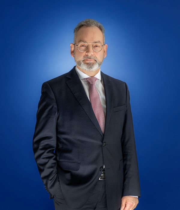 Das Bild zeigt Günther Paul vor einem blauen Hintergrund, der einen schwarzen Anzug trägt und erst in die Kamera schaut. Er trägt sein Sakkko zugeknöpft sowie eine rot-weiß gepunktete Krawatte und ein weißes Hemd. Seine rechte Hand steckt in der Hosentasche, sein linker Arm hängt locker an der Seite herunter.