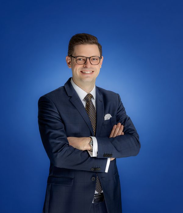Das Bild zeigt Sebastian Reffert. Er hat seine Arm vor der Brust verschränkt. Er steht vor einem blauen Hintergrund und lächelt in die Kamera.