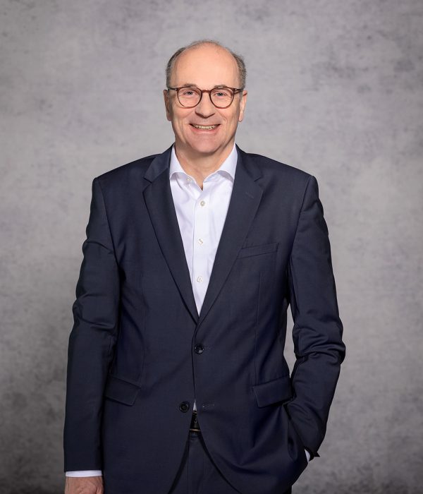 Im Bild ist Vorstandsmitglied der Volksbank Kassel Göttingen Hans Christian Reuß vor einem grauen Hintergrund zu sehen. Er trägt einen dunkelblauen Anzug sowie ein weißes Hemd. Sein Sakko ist zugeknöpft. Seine linke Hand steckt in der Hosentasche, sein rechter Arm wiederum hängt an der Seite herunter.