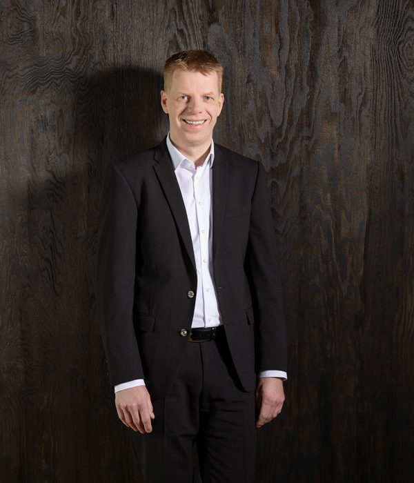 Das Bild zeigt Vorstandsmitglied Thorsten Schartel vor einer dunkelbraunen Holzwand. Er trägt einen schwarzen Anzug sowie ein weißes Hemd. Beide Arme hat er locker an der Seite herunterhängen und er lächelt fröhlich in die Kamera.