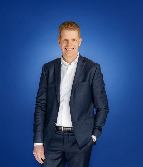 Das Bild zeigt Thorsten Schartel vor einem blauen Hintergrund. Er trägt einen dunkelblauen Anzug, sein Sakko ist offen, sodass sein weißes Hemd gut zu sehen ist. Seine linke Hand steckt in der Hosentasche. Sein rechter Arm wiederum hängt locker an der Seite herunter. Er lächelt fröhlich in die Kamera.
