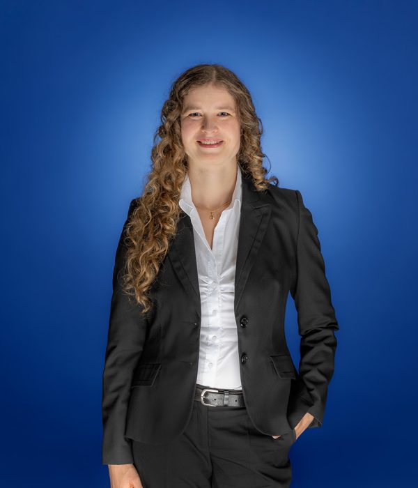 Das Bild zeigt Christine Schneider, die locker, vor einem blauen Hintergrund, in die Kamera lächelt.