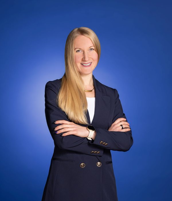 Das Bild zeigt Anna-Katharina Sögtrop. Sie steht vor einem blauen Hintergrund und hat ihre Arme vor der Brust verschränkt. Dabei lächelt sie in die Kamera.