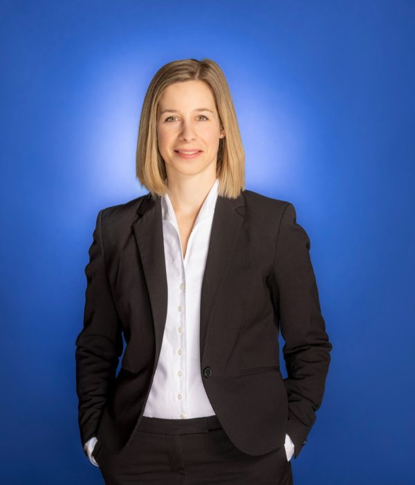 Das Bild zeigt Hanna Wenderoth, wie sie, vor einem blauen Hintergrund und beide Hände in ihre Hosentasche steckt, in die Kamera lächelt.