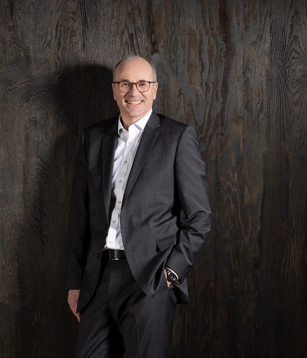 Das Bild zeigt Vorstandsmitglied Hans-Christian Reuß vor einer dunkelbraunen Holzwand. Er trägt einen dunkelgrauen Anzug und ein weißes Hemd. Seine linke Hand steckt in der Hosentasche, sein rechter Arm hängt locker an der Seite herunter. Er lächelt in die Kamera.
