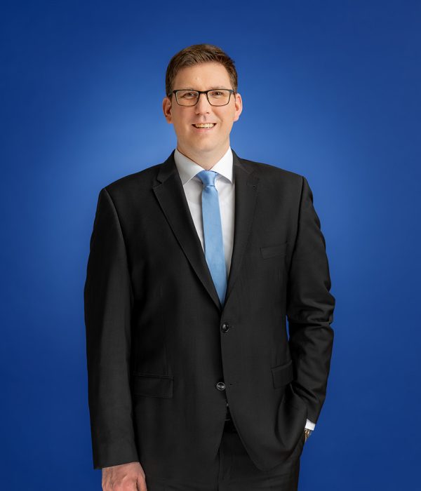 Das Bild zeigt Martin Hasselbein in einem schwarzen Anzug. Er trägt eine Brille und lächelt in die Kamera. Er trägt eine hellblaue Krawatte und ein weißes Hemd. Sein Sakko ist zugeknöpft. Seine linke Hand steckt in der linken Hosentasche, sein rechter Arm hängt locker an der Seite herunter.