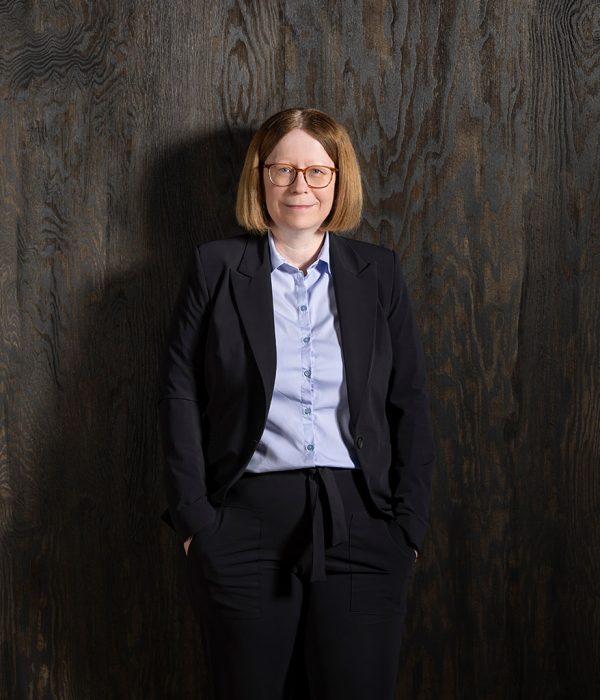 Das Bild zeigt Vorstandsmitglied Kerstin Orth vor einer dunkelbraunen Holzwand. Sie trägt einen schwarzen Anzug und eine hellblaue Bluse. Beide Hände stecken in der Hosentasche. Sie schaut schmunzelnd in die Kamera.